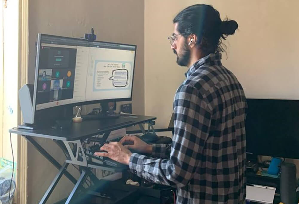 Arpan Kaul, on zoom, wearing a plaid shirt and earbuds, standing in front of his computer in his apartment, typing on his keyboard, teaching remotely.