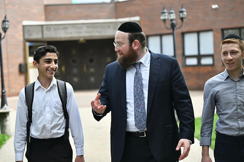  Rabbi Dovid Kurz, walking in front of school, talking with 2 male students, one on each side